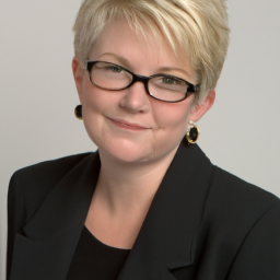 a headshot of a woman with short blonde hair and glasses, looking directly at the camera. she appears to be in her late 50s or early 60s, and is wearing a black blazer.