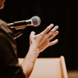 description: an anonymous image shows a person holding a microphone, passionately speaking at a podium in front of a diverse audience. the person's face is not visible, but their body language exudes confidence and determination.