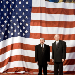 Two elderly men in suits standing in front of a large American flag.